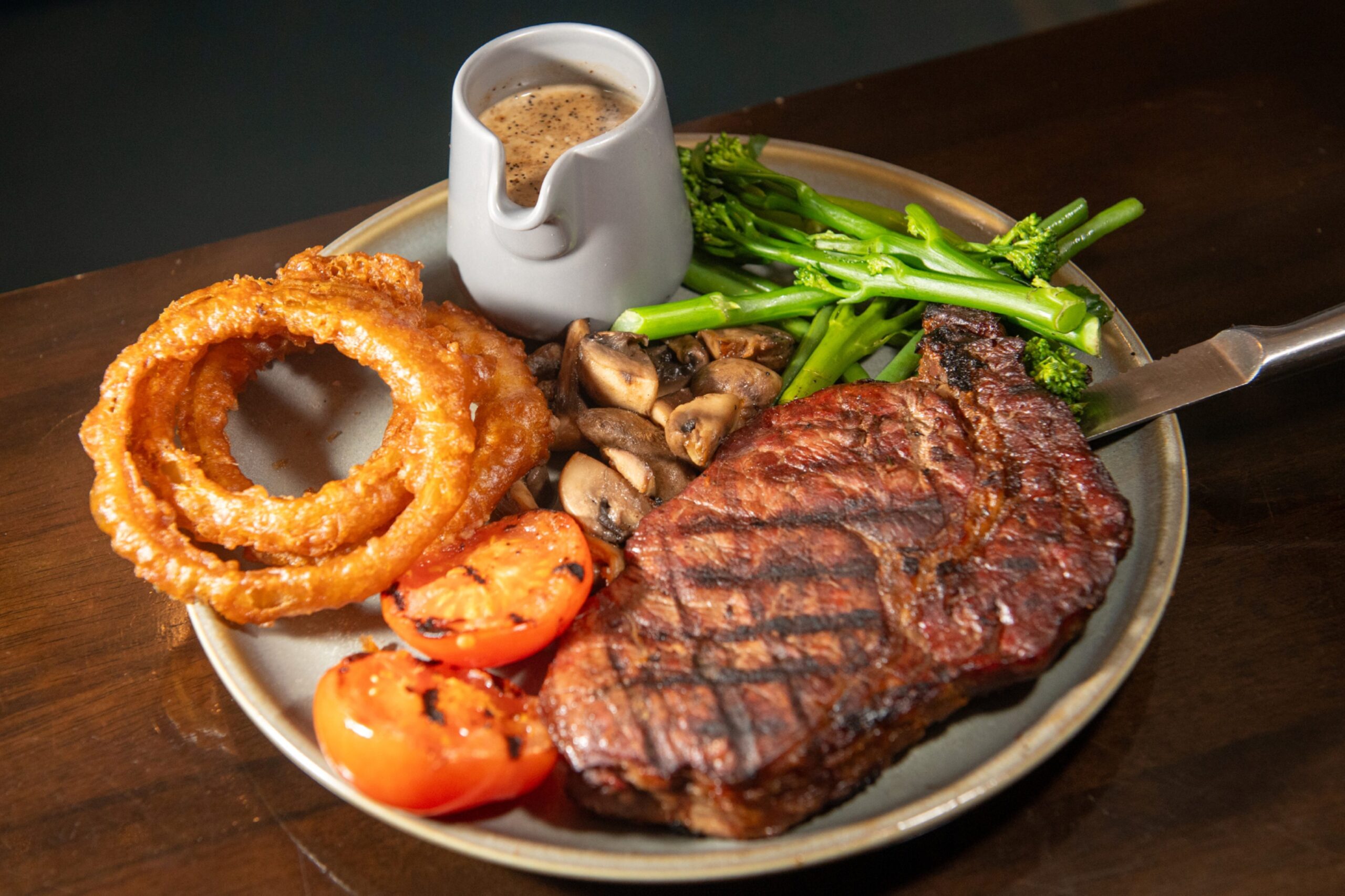 A steak with long stem broccoli, onion rings, mushrooms, tomato and a jug of sauce