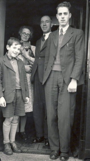 A young Kenny Duffus in short trousers pictured in the doorway of his home with his elder brother and dad in suits, and mother - holding a cat - in the background.