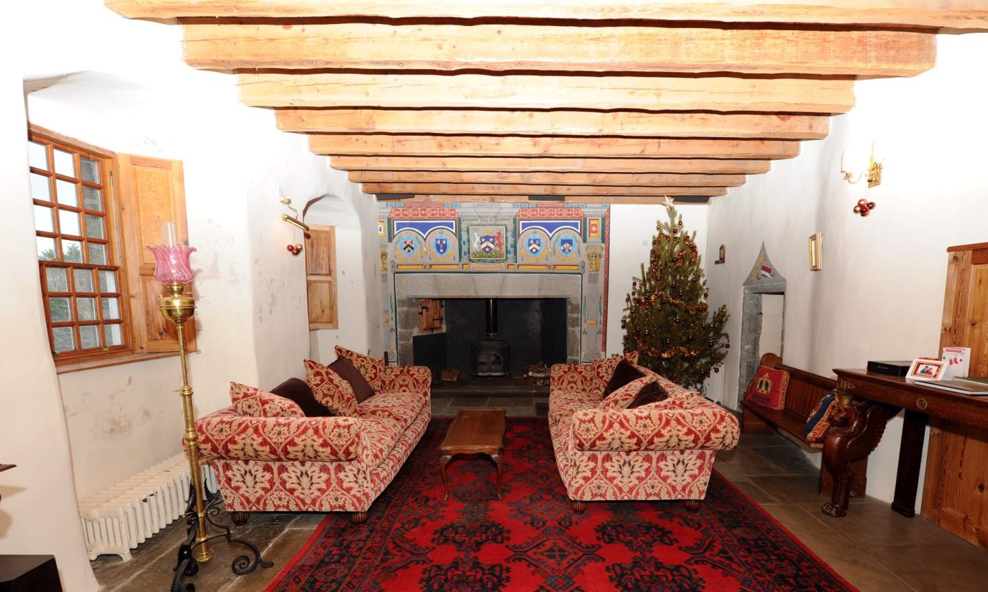 Inside a living area of the abode, with red and gold patterned sofas, a wooden coffee table, a wooden bench and a stone fireplace with clan crests above it