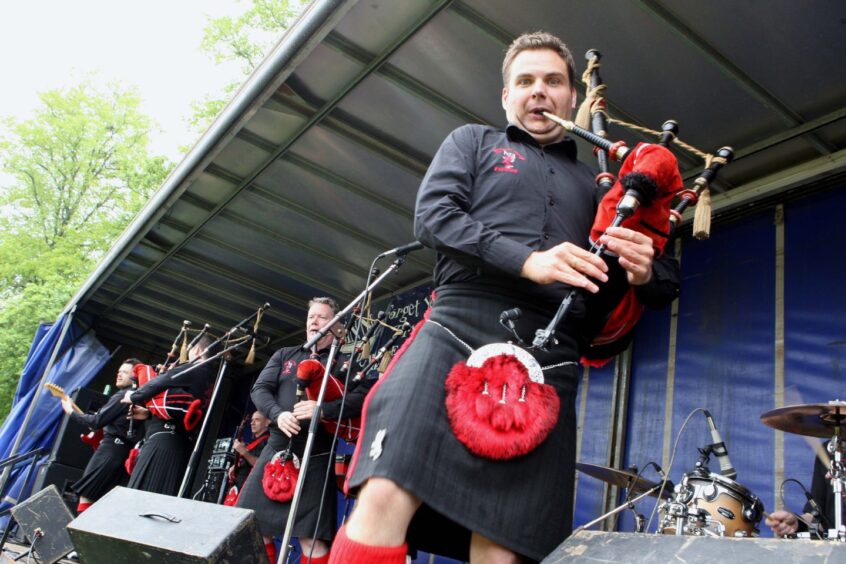 Stuart looks directly into the camera while playing the bagpipes on stage in Perth. He is dressed in a kilt.