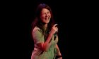 Comedian Rosie Jones with a mic laughing on a black background.