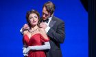 Elly Jay smiling in a red dress playing Vivian Ward in Pretty Woman the Musical while her co-star playing Edward places a necklace on her.