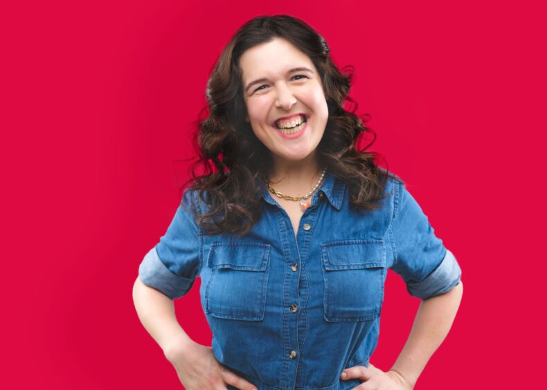 Rosie Jones smiling at the camera on a red background in a denim jumpsuit.