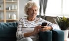 Happy smiling older woman using phone, looking at screen, sitting on cozy sofa in living room, excited mature senior female chatting in social network or shopping online, having fun with smartphone.