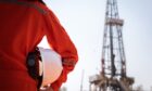 A worker is holding safety hardhat or helmet with blurred background of drilling rig derrick structure.
