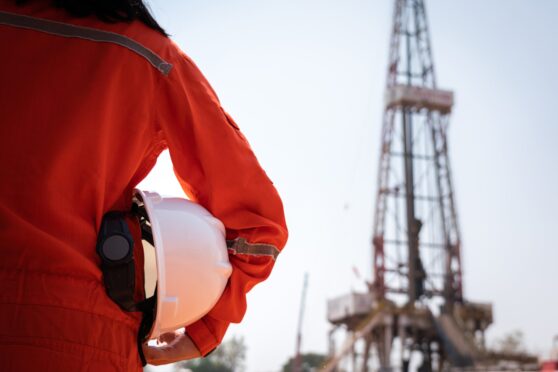 A worker is holding safety hardhat or helmet with blurred background of drilling rig derrick structure.