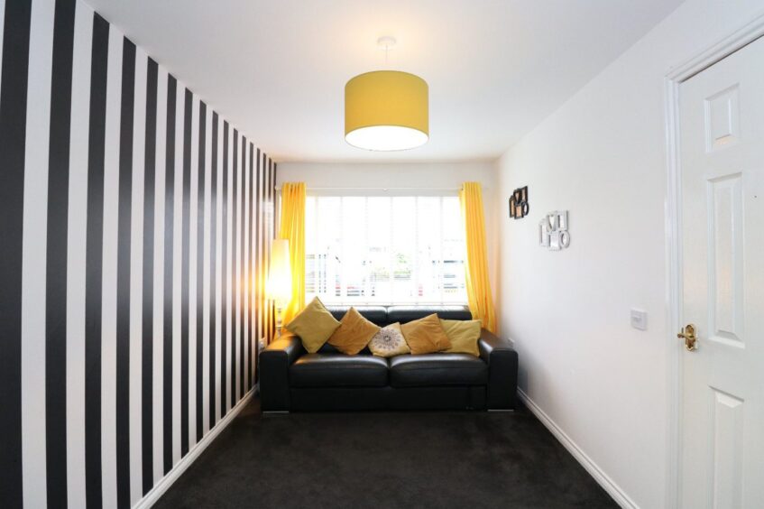 A room with a black carpet and sofa, a black and white striped accent wall and yellow cushions, light shade and curtains