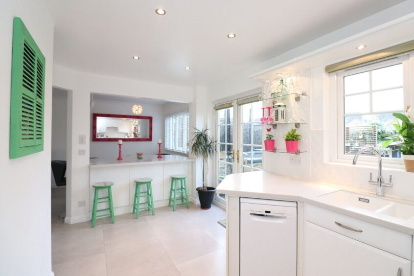 The open plan kitchen and dining area in the Kintore home after the renovation. The walls, counters, cupboards and floor tiles are white while the small accents like bar stools, mirror frame, candle holders and plant pots are mint green and bright pink