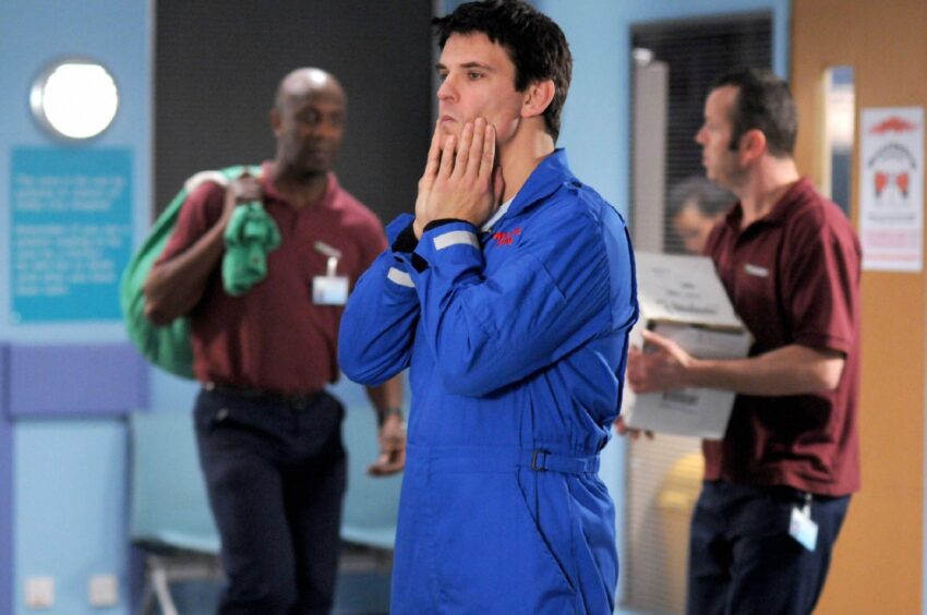 Tristan Gemmill playing Adam in Casualty holding his cheeks with both hands in a hospital. 