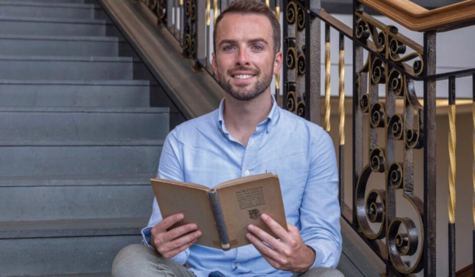 Shane Strachan holding an open book and smiling at the camera. 