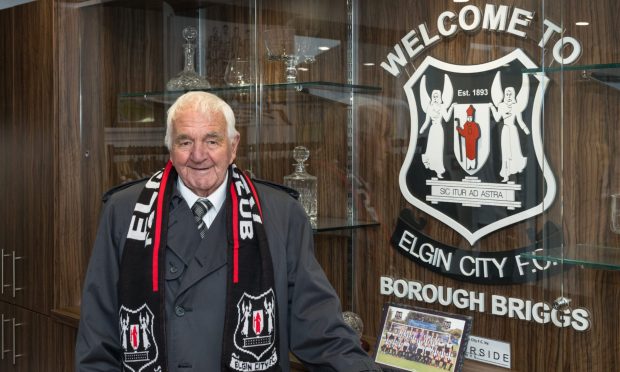 John Blackhall wearing Elgin City scarf in front of club sign