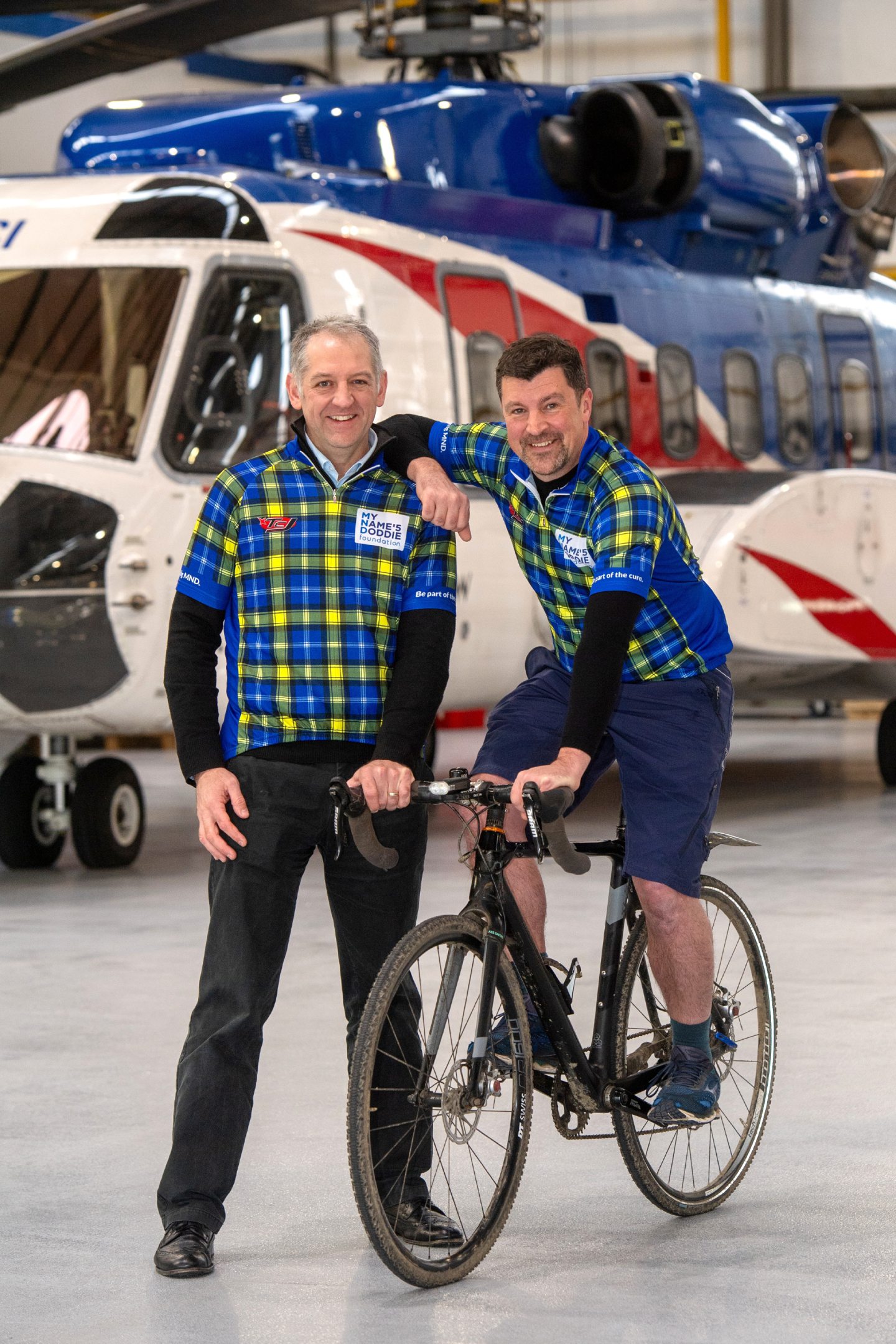 John Fyall sits on a bike and leans on Michael Fotheringham at Aberdeen Airport