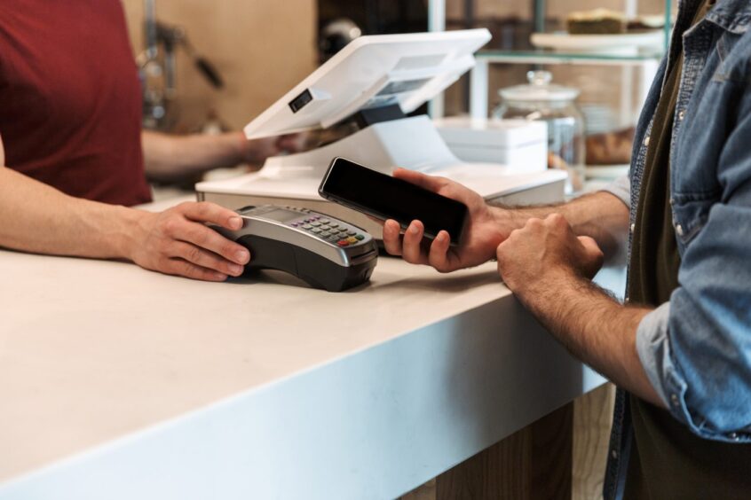 Cropped photo of young man wearing denim shirt paying debit card in cafe, while waiter holds payment terminal.