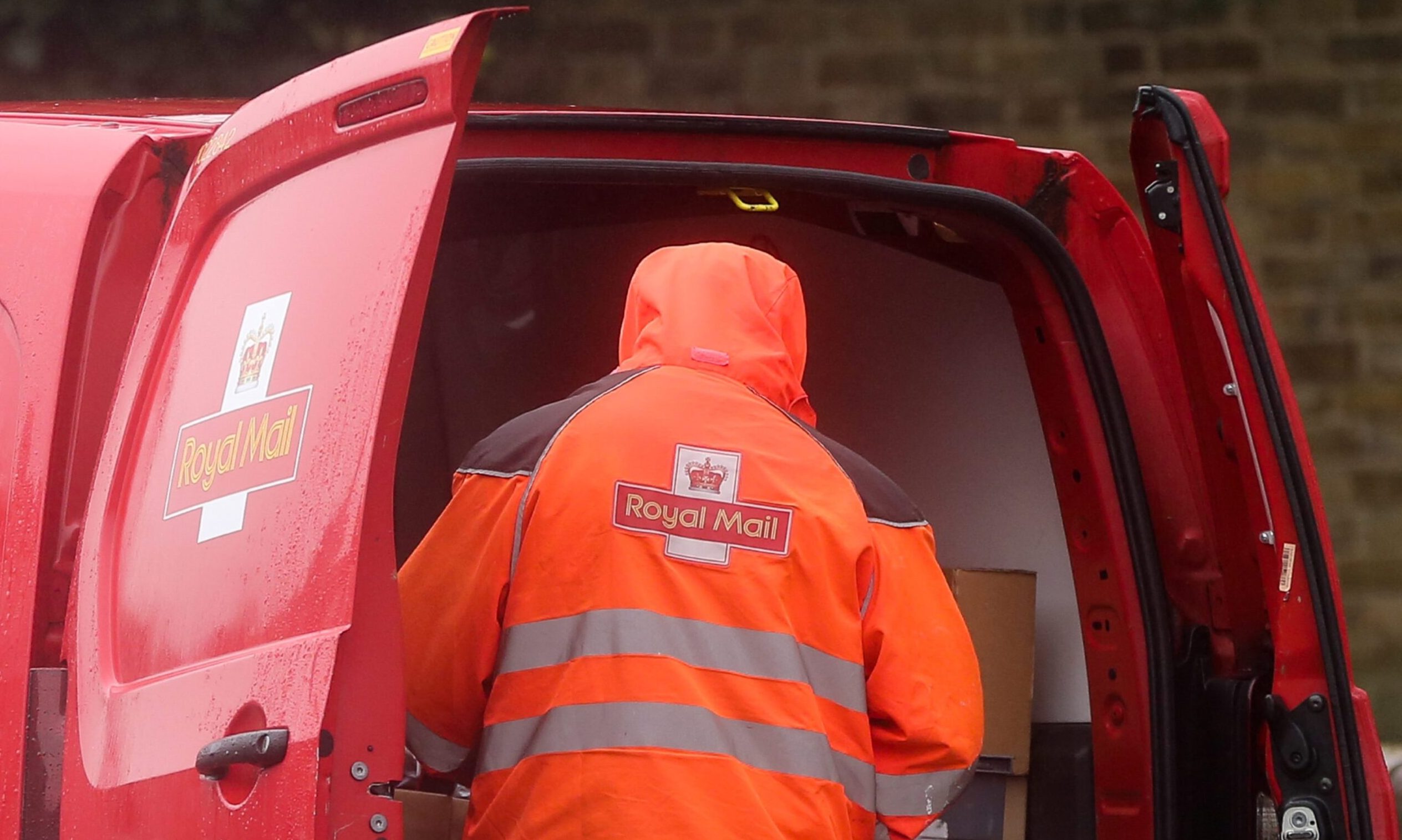Royal Mail worker wearing an orange jacket at the rear of a red Royal Mail van with the doors open.