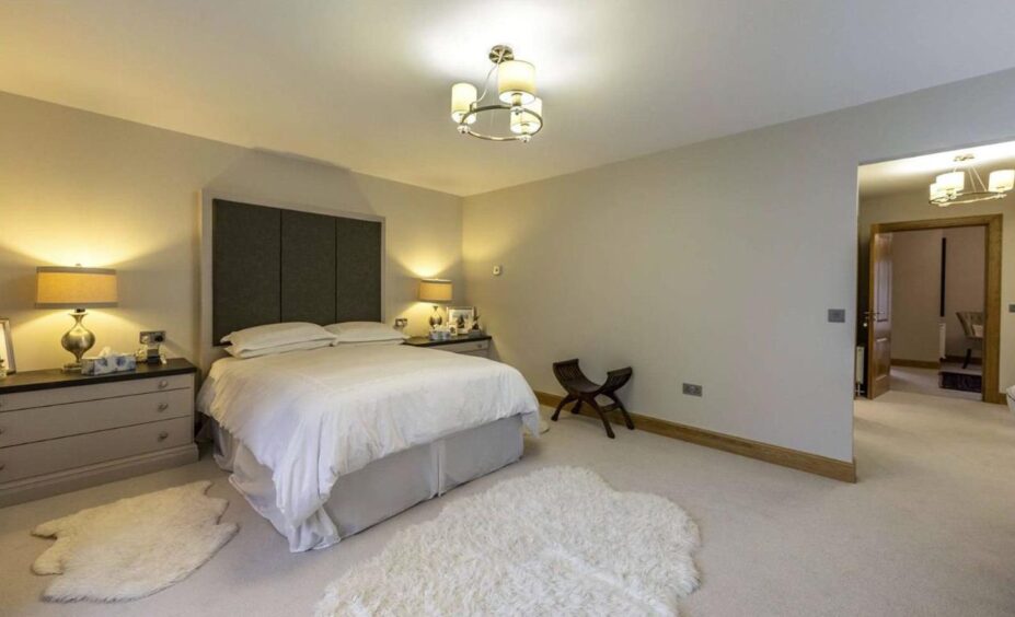 The master bedroom, with neutral tones. It has a double bed, two shaggy white rugs and two sets of drawers with bedside lamps on them 