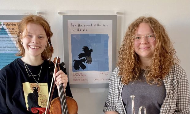 Two young women smile, holding instruments, in front of a poster for their play For the Sea and Sky.