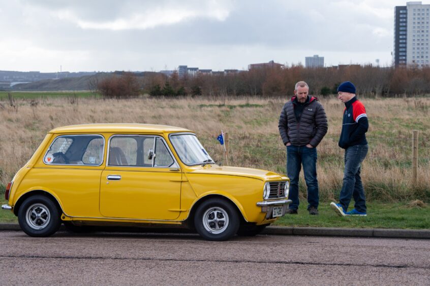 Mini enthusiasts stand on the grass verge near a yellow mini.