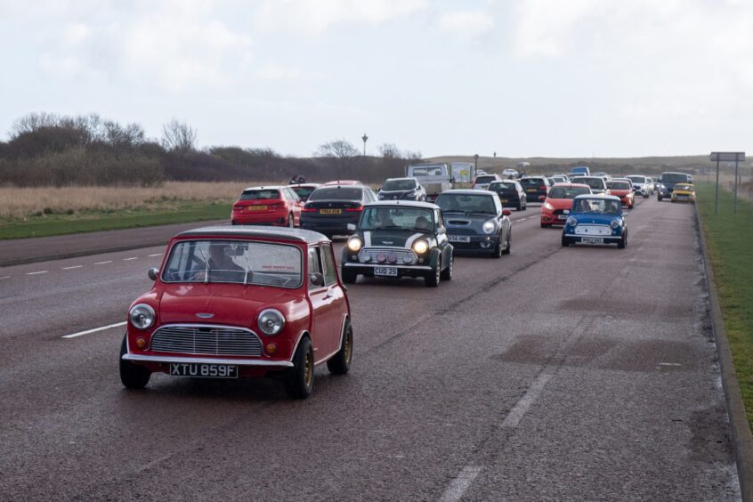 Mini's of all models and colours drive down a busy road.