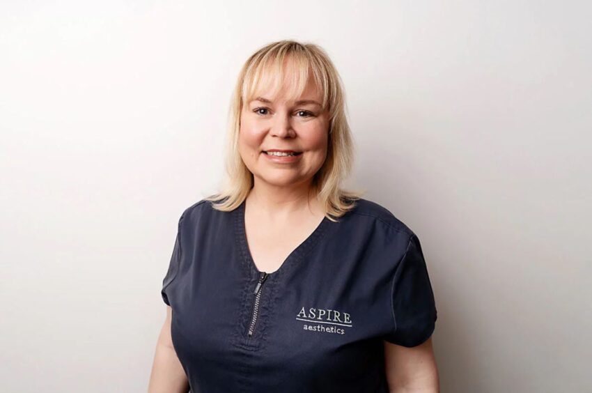 Linda Strachan from Aspire Aesthetics. She is smiling standing in front of a white wall. 