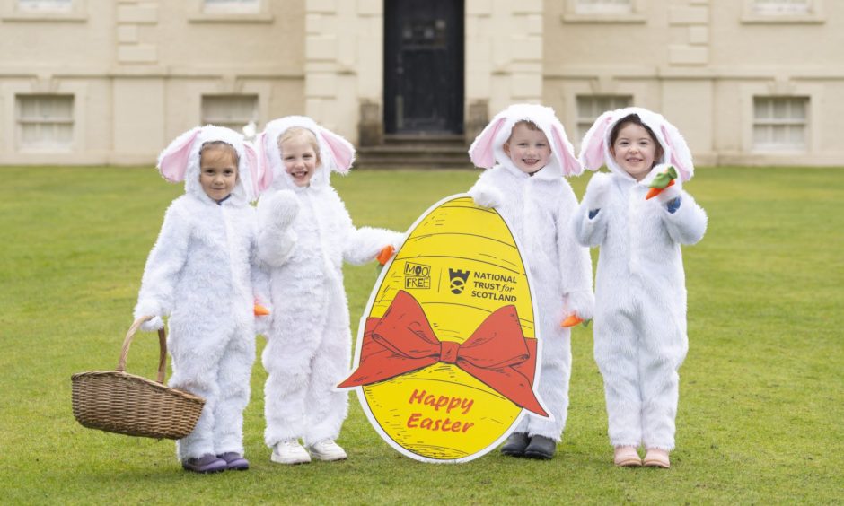 Four children in bunny costumes holding up a cardboard Easter egg