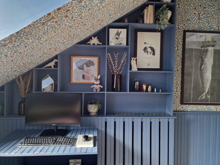 The dusty blue shelving unit in the smallest bedroom. There is a small desk with a computer on it and lots of knickknacks on the shelves along with some framed artwork