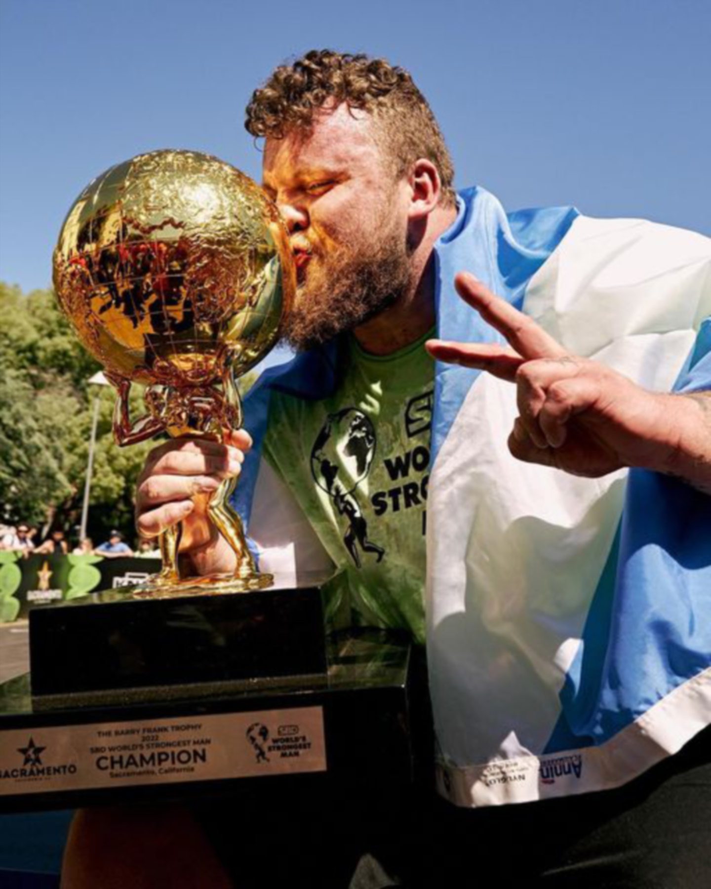 Tom Stoltman kissing Worlds Strongest Man trophy with Scotland flag wrapped round him. 