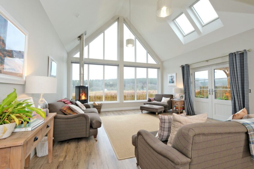 A living area with lots of natural light coming through a large set of windows along one wall. There are double doors leading outside and a standalone woodburner in the corner, surrounded by sofas and armchairs.