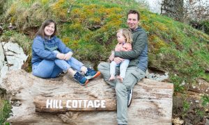 Kayleigh and Jamie Macleod sitting on a wooden sign saying hill cottage with their daughter Bella.