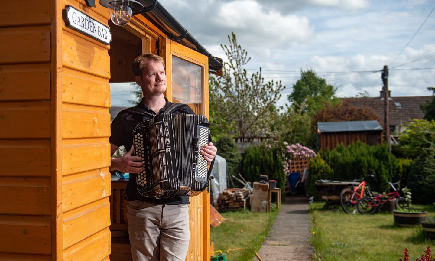 Graeme Mackay outside shed in back garden playing accordion. 