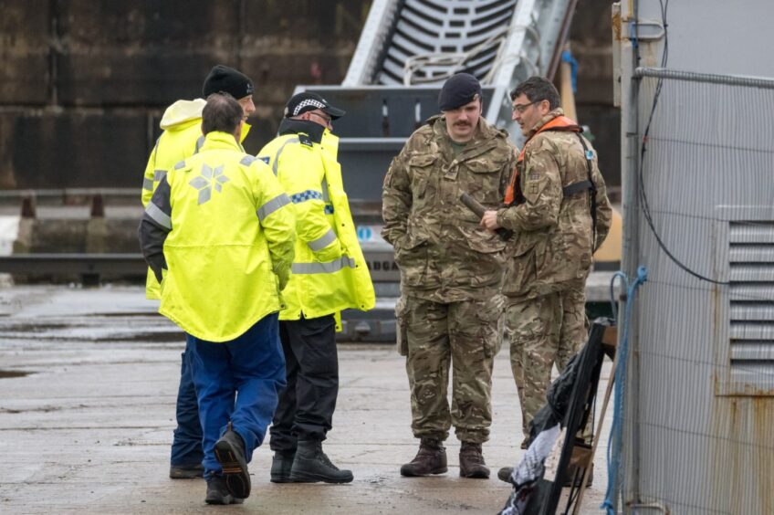 Individuals dressed in military uniform and fluorescent yellow coats stand at the pier.