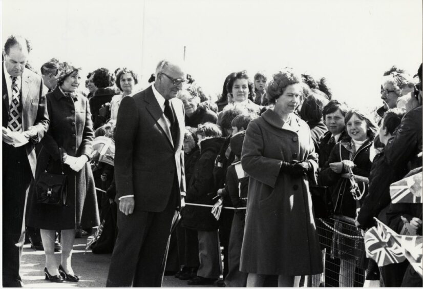 Queen Elizabeth walks alongside crowds including many children. She is accompanied by dignitaries.