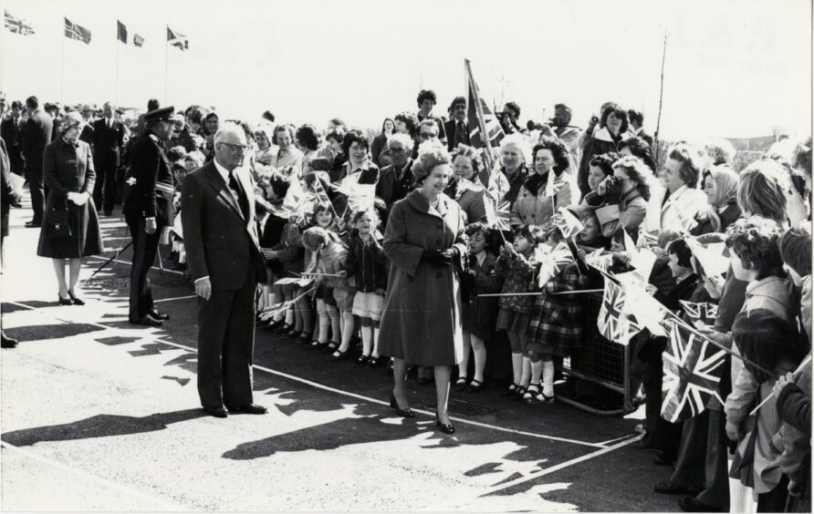 A smiling Queen meets the people during her walkabout at the St Fergus terminal. Also in picture is Monsieur Rene Granier de Lilliac, chairman of Total Oil. Many children waving flags are present.