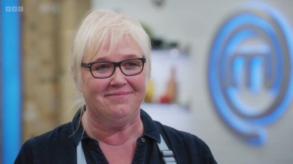 Muir with blonde hair wearing a blue blouse and apron with black glasses. 
