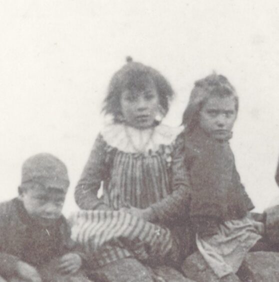 A close up of Mary Ann Mackenzie sitting in between two other children. She is about 11 years old and wearing a striped pinafore. Image from Ullapool Museum.
