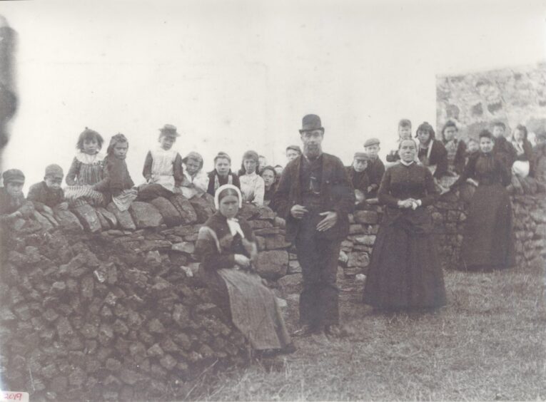 A black and white picture of the pupils and staff of Scoraig primary school, 1895. The children are arranged on top of and along a wall, while four staff, identities unknown, dominate the foreground, along with a peat stack. Image supplied by Ullapool Museum.