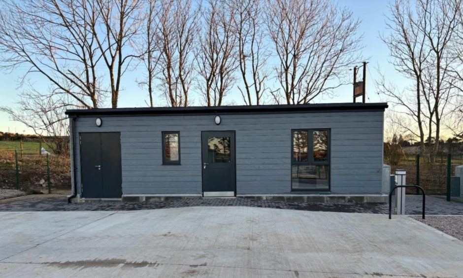 Grey building with black roof in front of a forest.