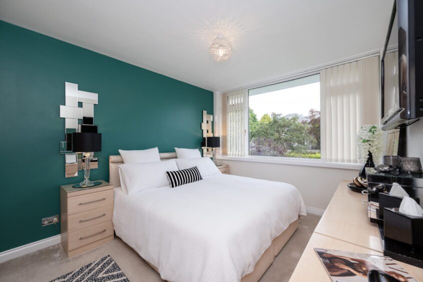 A bedroom in the home with a jade green accent wall, a double bed, side tables and lamps, statement mirrors and a wall-mounted television above a unit