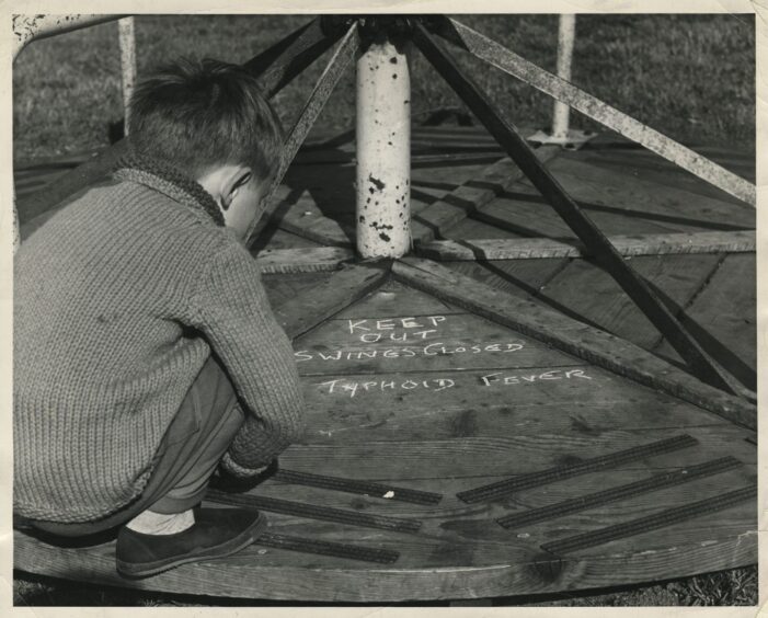 A small boy looks at the upturned roundabout in his playground bearing the words: "KEEP OUT. SWINGS CLOSED. TYPHOID FEVER."