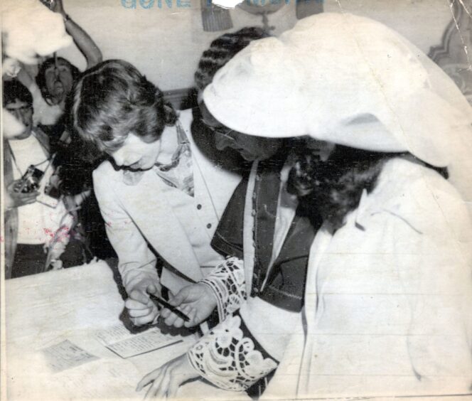 Mick with a pen in his hand looking at the registry, flanked by the priest and Bianca in the foreground.