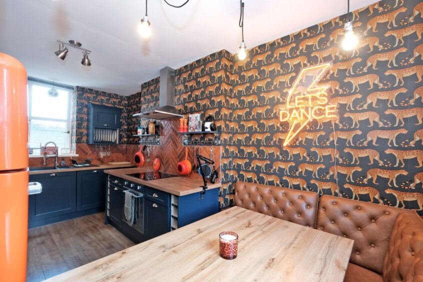 The kitchen and dining room with leopard wallpaper, a yellow neon sign, navy cupboards with wooden countertops and burnt orange booth seating