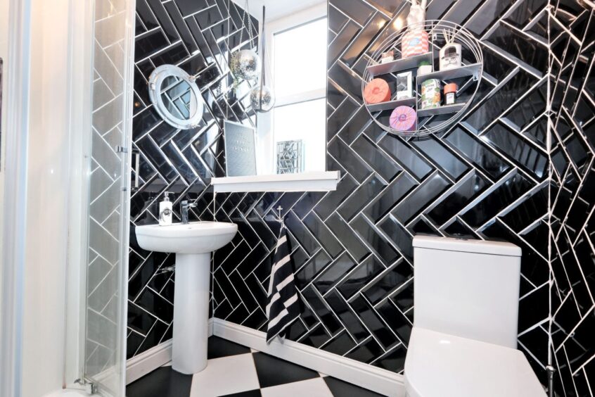 The bathroom with black and white floor tiles, glossy black herringbone wall tiles, matching white toilet and sink and a cubical shower