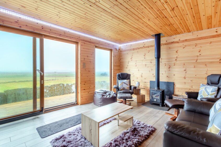 Another view inside the summer house at the Thurso property, with sofa, a freestanding woodburning stove and large windows with views of the Morven hills.