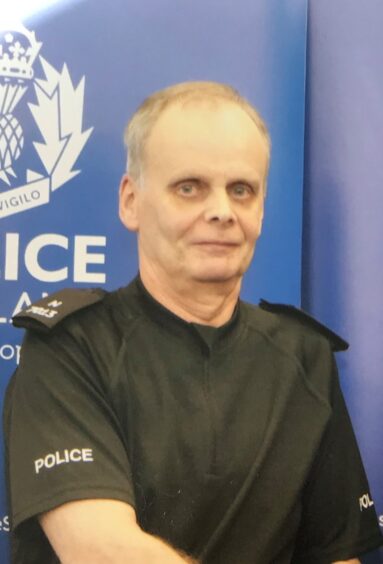 Iain Sutherland dressed in a black police uniform standing in front of a blue police sign.