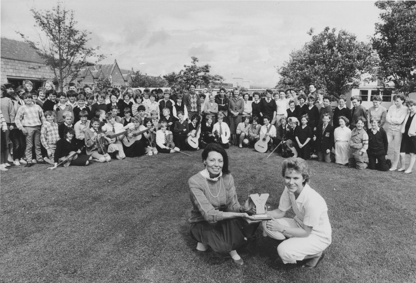 Two students with a trophy sitting on the grass, surrounded by a large group of pupils