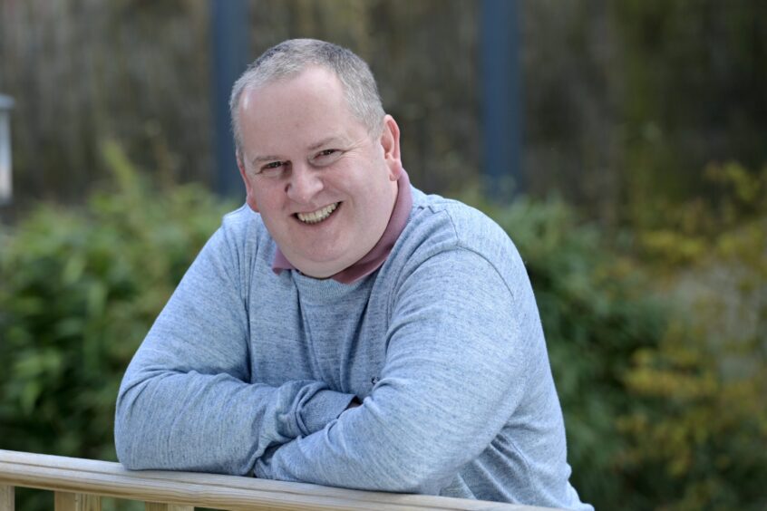 Glenn dressed in a light blue jumper leaning against a fence post.