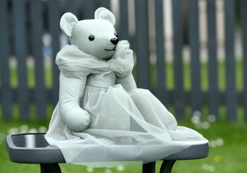 Memory bear wearing a dress sitting on a glass table outdoors with grass and a brown fence in the background.