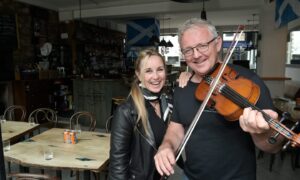 A blonde woman and a grey-haired man holding a fiddle looking at the camera standing side-by-side in a pub.