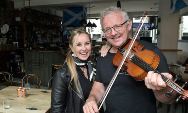 A blonde woman and a grey-haired man holding a fiddle looking at the camera standing side-by-side in a pub.