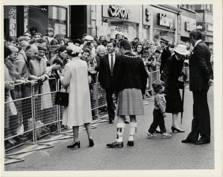 Three year old Andrew Bell, 26 Broom Dive, Inverness, who cross the High Street to present the Queen with flowers, is escorted back to his granny by an  equerry and the Queen's Lady-in-Waiting."