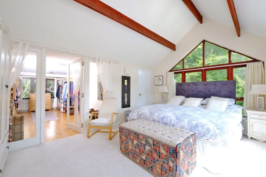 One of the bedrooms with a large set of windows behind the double bed, the walls and carpet a white and there are french glass doors leading to another room with rails of clothing in it